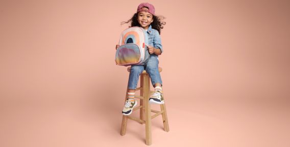 Ett leende barn sitter på en hög träpall mot en beige bakgrund. Barnet bär jeanskläder, en rosa keps och håller i en färgglad ryggsäck med regnbågsmotiv.