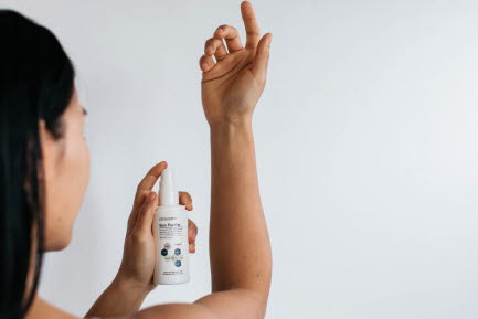 En person med l&aring;ngt, m&ouml;rkt h&aring;r sprayar en produkt fr&aring;n en vit flaska p&aring; sin arm mot en ljus bakgrund.