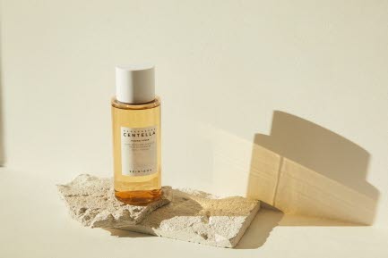 En flaska med Skin1004 Centella hudv&aring;rdsprodukt st&aring;r p&aring; ett stenblock, framf&ouml;r en ljus beige bakgrund. Flaskan har en enkel etikett med vit text.